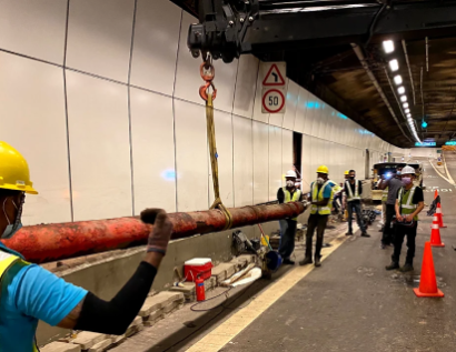 Workers in safety gear guide a crane lifting a large red pipe inside a tunnel, ensuring proper alignment and installation.